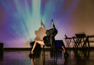 Image: A photograph taken in Salt Lake City during a work-in-progress showing of "I'll Never Be Beyoncé" for loveDANCEmore's Sunday Series performance platform, November 2022. Alexandra, wearing a black mesh top, sequined bottoms, and black high heel shoes straddles a black folding chair. Her back is facing the audience. She is looking up singing into a microphone. Upstage is a backdrop with hues of purple and blue fading into each other. Photograph by Todd Collins.